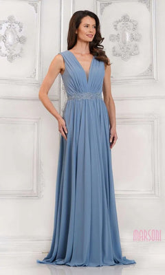 A woman in the Marsoni MV1292 long formal dress, featuring a sleeveless, floor-length light blue chiffon design with V-neckline and ruched bodice, stands against an ornate white wall.