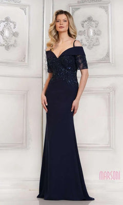 A woman in the Marsoni MV1295 long formal dress in navy, featuring beaded embellishments, stands before an ornate white wall.