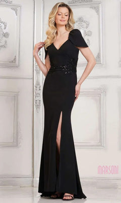 A woman stands against an ornate white wall in the Marsoni MV1297, a black floor-length crepe sheath dress with short sleeves, V-neckline, side slit, and beaded waist detail.