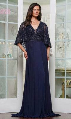 A woman wearing the Marsoni MV1302 long formal dress in navy, featuring beaded details and a sheer cape overlay, stands before white French doors.
