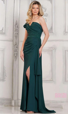 A woman models the Marsoni MV1304 long formal dress in deep green, a floor-length, one-shoulder gown with a thigh-high slit, posing against an ornate white wall.