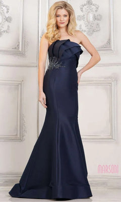 A woman stands before a decorative white wall in the navy Marsoni MV1308, a strapless mermaid long formal dress adorned with floral embellishments, showcasing its elegant design.
