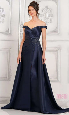 A woman wears the Marsoni MV1310 long formal dress, an off-the-shoulder navy blue gown with beaded embellishments and a mermaid silhouette, standing before an ornate white wall.