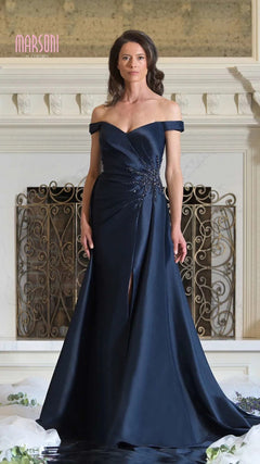 A woman stands indoors in the Marsoni MV1310 long formal dress, a navy off-the-shoulder gown with a mermaid silhouette, front slit, and embellishments, posed before an ornate fireplace.