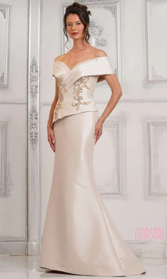 A woman wears the Marsoni MV1312 long formal dress in champagne, featuring an off-shoulder fitted stretch Mikado design with embroidered details and a mermaid skirt, posing before an ornate, light-colored wall.