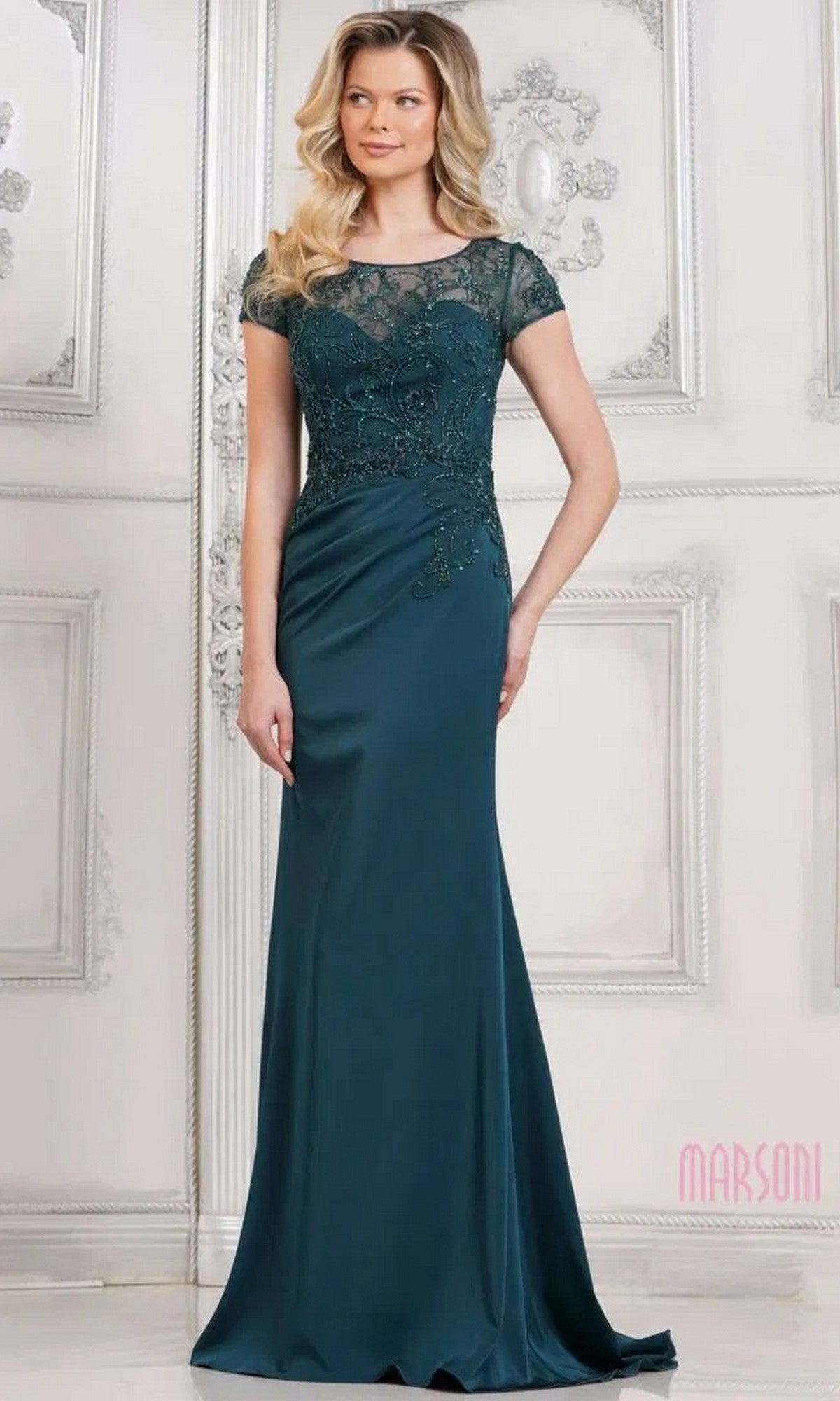 A woman in the Marsoni MV1317 long formal dress in deep green, featuring a sheath skirt, beaded bodice, and short sheer sleeves, stands before an ornate white wall.