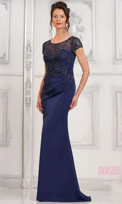 A woman wearing the Marsoni MV1317 Long Formal Dress in navy, featuring a beaded-embellished bodice and short sleeves, stands against an ornate white wall.