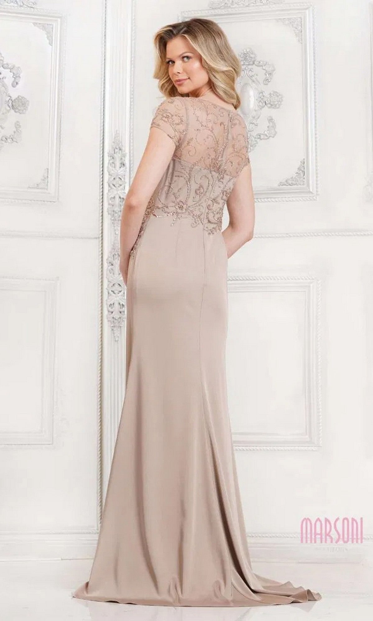 A woman stands facing away in an elegant, white-decorated room wearing the Marsoni MV1317 Long Formal Dress, featuring a beaded-embellished bodice and sheer, embroidered short sleeves.