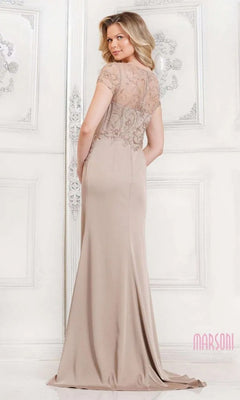 A woman stands facing away in an elegant, white-decorated room wearing the Marsoni MV1317 Long Formal Dress, featuring a beaded-embellished bodice and sheer, embroidered short sleeves.