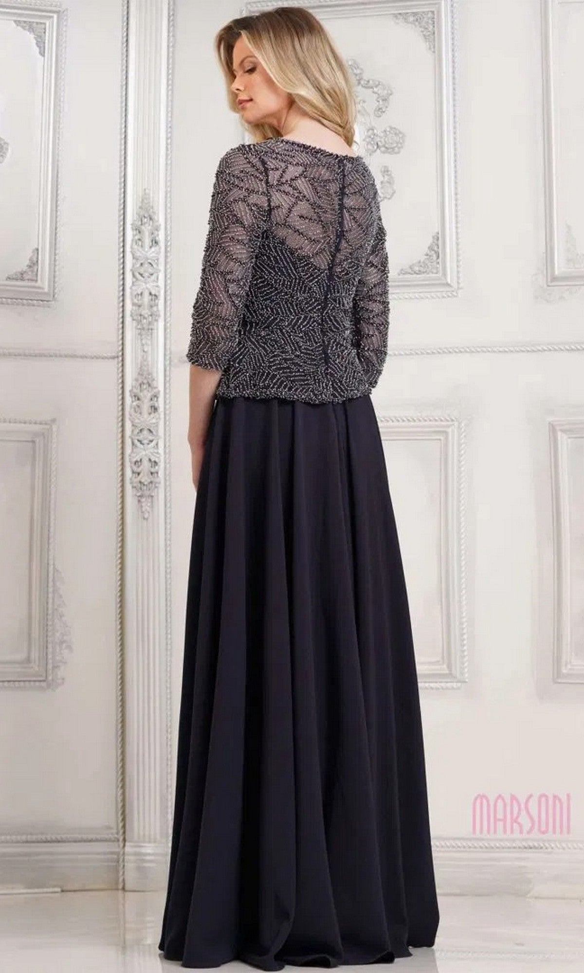 A woman stands in an elegant room wearing the Marsoni MV1319 long formal dress, featuring a floor-length black A-line skirt and a patterned, semi-sheer top with three-quarter sleeves.