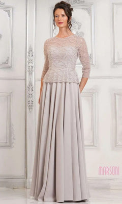 A woman wears the Marsoni MV1319 long formal dress in oatmeal, featuring a beaded bodice with three-quarter sleeves and an elegant A-line skirt, standing against an ornate white wall.
