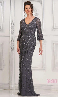 A woman stands before a white ornate wall in the Marsoni MV1320 long formal dress in charcoal, featuring three-quarter sleeves and silver floral embellishments.