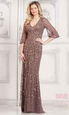 A woman stands before a decorative wall, wearing the Marsoni MV1320 long formal dress in dark taupe with sheer sleeves and floral embroidery—a refined, modest gown ideal for any elegant event.