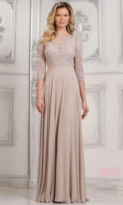A woman in the Marsoni MV1322 long formal dress in champagne, featuring a lace embroidered bodice and three-quarter sleeves, stands against an ornate, light-colored wall.
