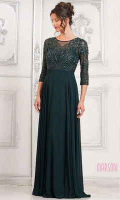 A woman stands in an elegant, white-paneled room wearing the Marsoni MV1322—a modest, floor-length formal dress in deep green with a lace embroidered bodice and three-quarter sleeves.