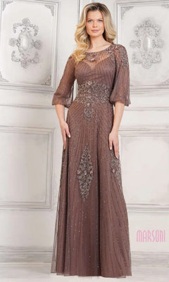 A woman in a floor-length, dark taupe Long Formal Dress: Marsoni MV1323 with sheer sleeves and intricate beadwork stands against an ornate, light-colored wall.