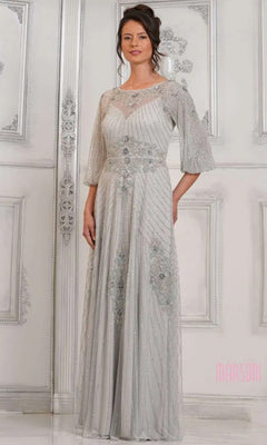 A woman in a silver Marsoni MV1323 long formal dress with sheer sleeves and intricate beading stands before an ornate white wall.