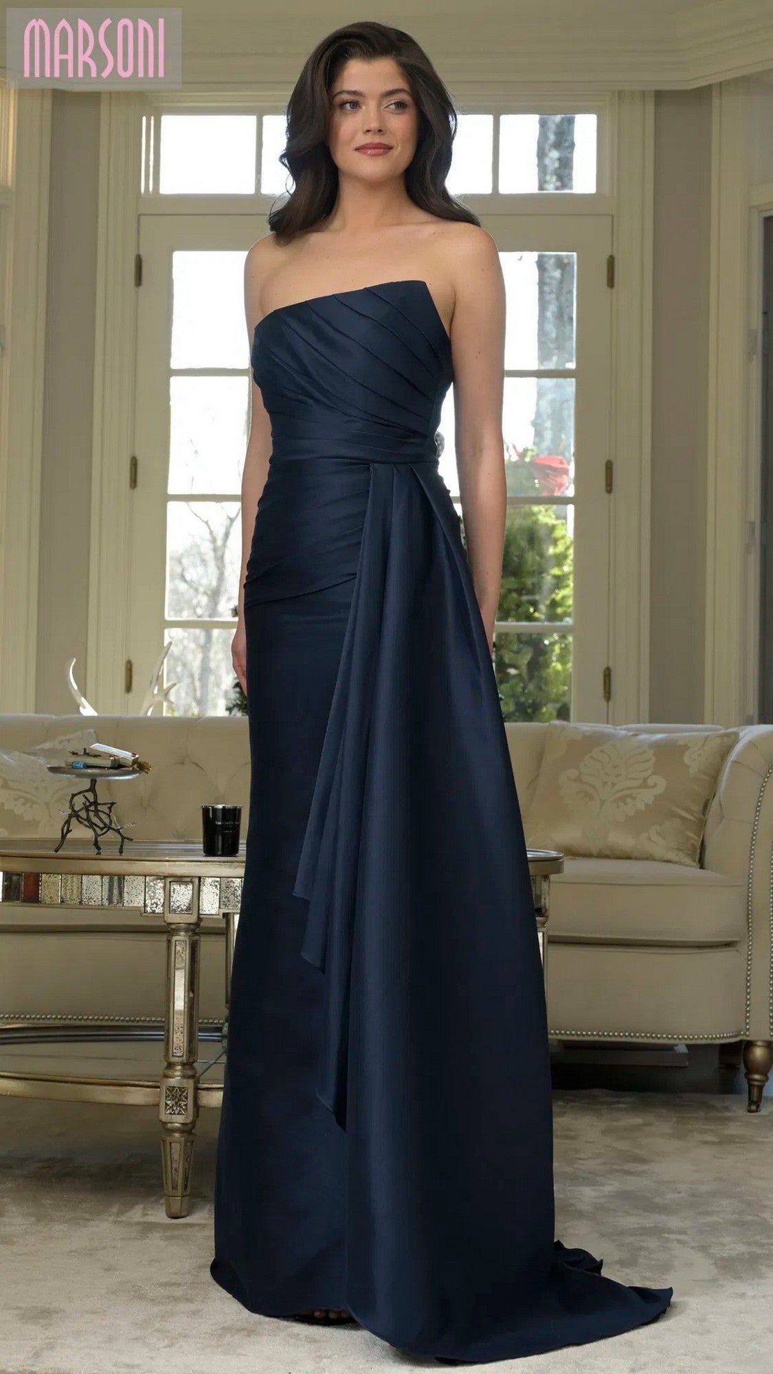A woman poses indoors in front of a beige sofa and large windows, wearing the Marsoni MV1326 long formal dress in navy with strapless design and elegant draped detailing.