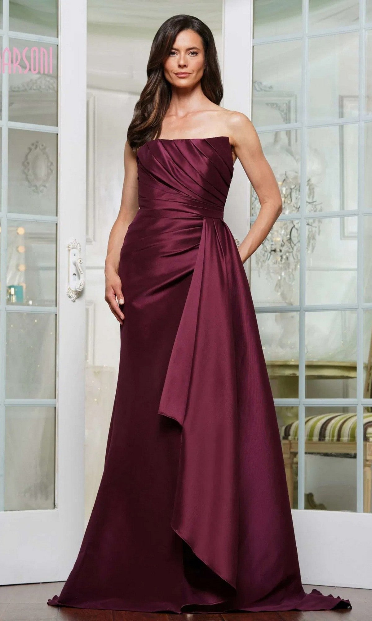 A woman stands in front of glass doors wearing the Marsoni MV1326 long formal dress in burgundy, featuring a sleeveless, floor-length silhouette with a draped, asymmetrical design.
