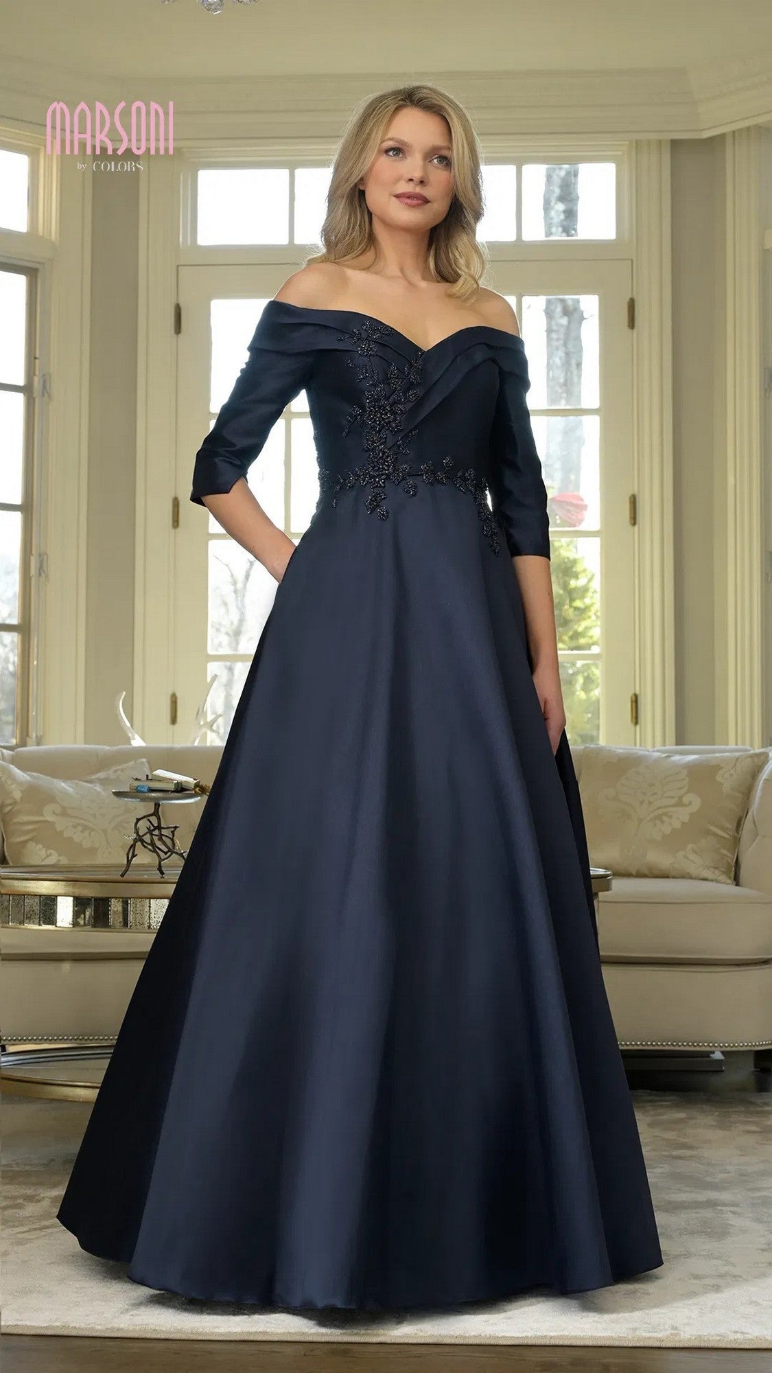 A woman poses gracefully indoors in front of large windows, wearing the Marsoni MV1336—a long formal navy dress with off-the-shoulder sleeves and floral embroidery.