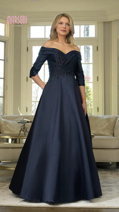 A woman poses gracefully indoors in front of large windows, wearing the Marsoni MV1336—a long formal navy dress with off-the-shoulder sleeves and floral embroidery.