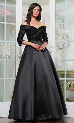 A woman stands indoors wearing the Long Formal Dress: Marsoni MV1336 in black—an off-the-shoulder, floor-length gown with three-quarter sleeves and floral appliqué on the bodice.