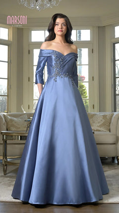 A woman stands in a well-lit living room wearing the Marsoni MV1336 long formal dress in slate blue, featuring off-the-shoulder satin fabric with floral embellishments.