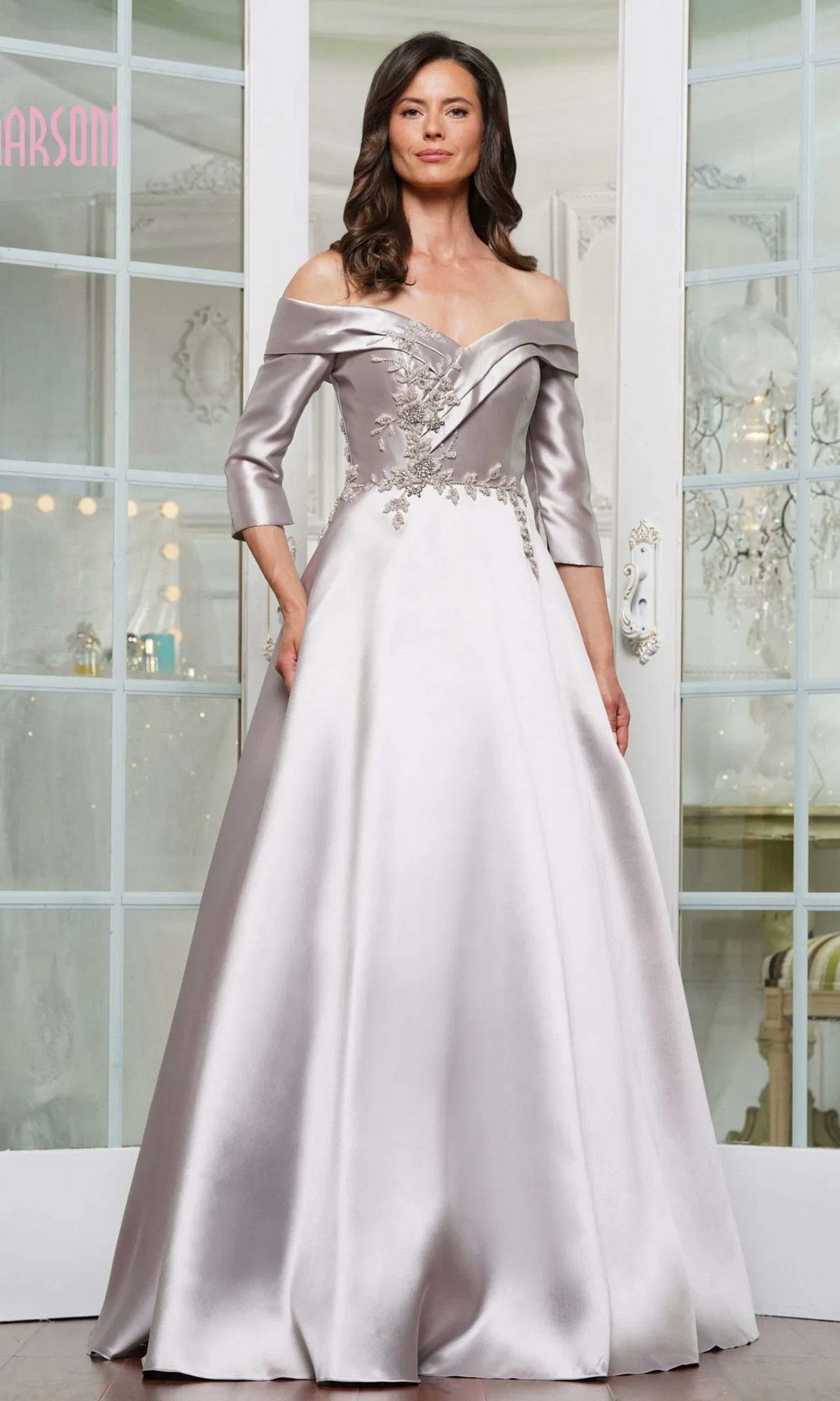 A woman in a taupe off-the-shoulder Marsoni MV1336 satin gown with floral embroidery stands indoors before glass doors.