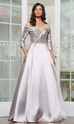 A woman in a taupe off-the-shoulder Marsoni MV1336 satin gown with floral embroidery stands indoors before glass doors.