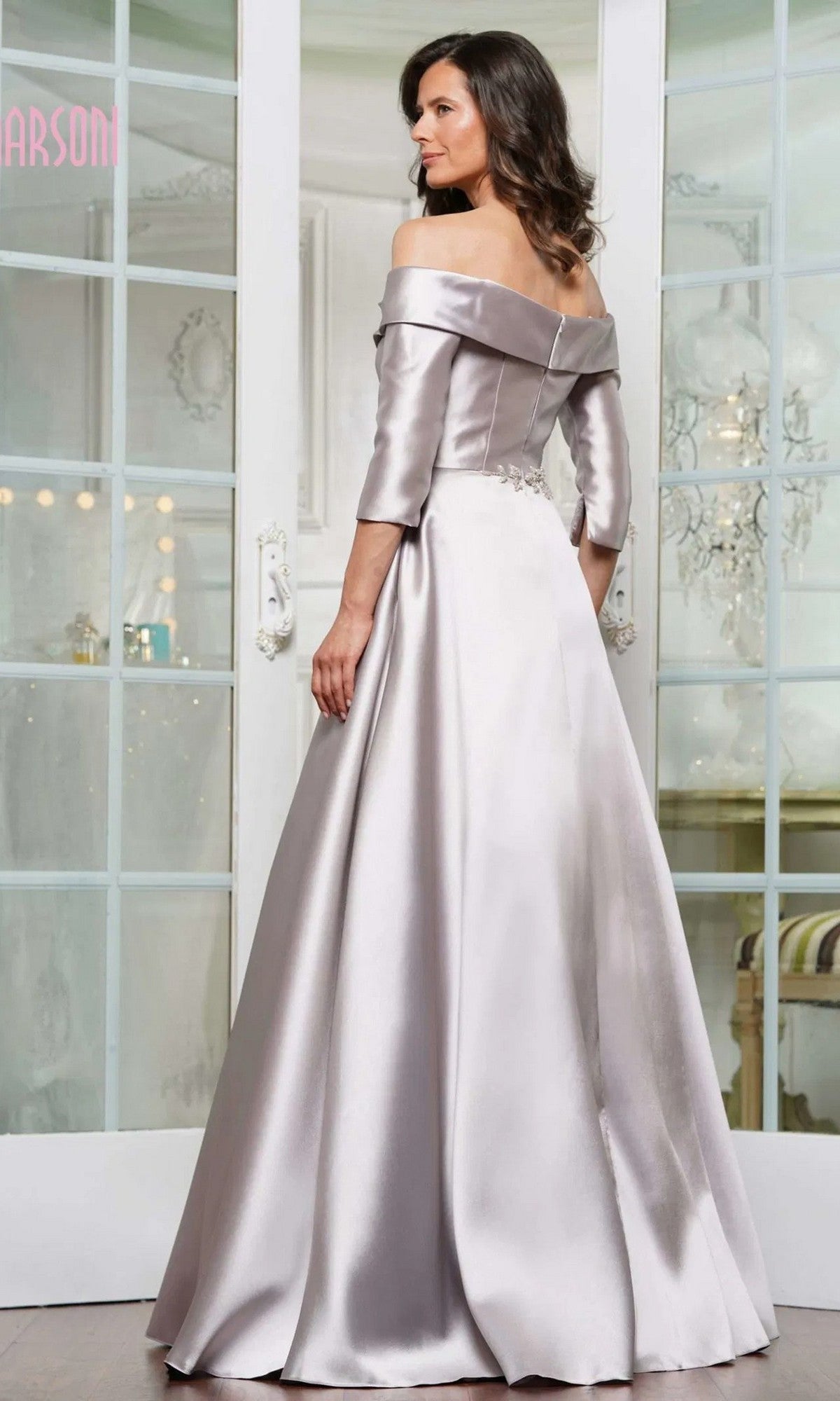 A woman in a floor-length, off-the-shoulder silver satin Marsoni MV1336 formal dress with three-quarter sleeves stands facing away before glass double doors.