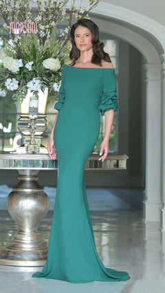 A woman stands indoors in a fitted, emerald Marsoni MV1337 long formal dress with ruched off-the-shoulder sleeves, posed beside a table topped with a tall floral arrangement.