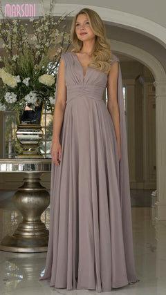 A woman stands indoors wearing the Marsoni MV1349 long formal dress in dusty rose, featuring chiffon fabric, long cape sleeves, draped shoulders, and a floor-length design. She poses before a table with a large floral arrangement.
