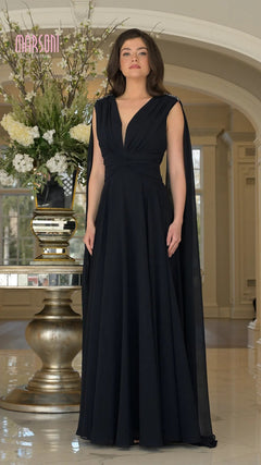 A woman stands indoors in front of a large floral arrangement wearing the Marsoni MV1349 long formal dress in midnight, featuring black chiffon fabric, long cape sleeves, a deep V-neck, and a ruched bodice.