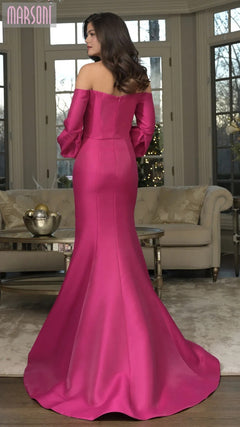 A woman wears the Marsoni MV1355 long formal dress with an off-the-shoulder mermaid silhouette, standing in a living room with neutral furniture and large windows.