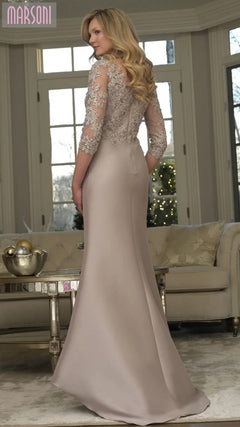 A woman in the Marsoni MV1357 long formal dress with lace sleeves stands in an elegant living room featuring large windows and neutral decor.