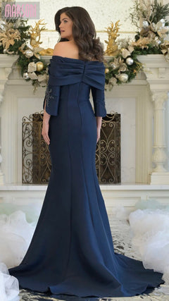 A woman stands facing away in front of a decorated white fireplace, wearing the Marsoni MV1360 long formal navy off-the-shoulder evening dress.