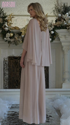 A woman in the Marsoni MV1361 long formal dress, featuring flowing light pink fabric and cape sleeves, stands with her back to the camera before a holiday-decorated fireplace.