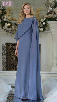 A woman in the Marsoni MV1362 long formal dress in slate blue with cape sleeves and crystal-beaded embellishment stands before a white fireplace decorated with greenery and ornaments.
