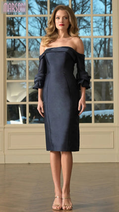 A woman stands indoors wearing the Marsoni MV1370 Knee Length Semi-Formal Dress in navy with ruffled off-the-shoulder sleeves and nude heels. Large windows behind her reveal a view of trees.