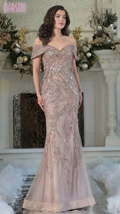 A woman stands indoors wearing the Marsoni MV1372 Long Formal Dress in taupe, an off-shoulder mermaid gown highlighted by intricate beaded embroidery. Floral decorations adorn the background.