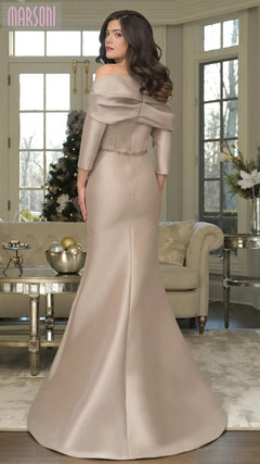 A woman in the Marsoni MV1374 long formal dress stands in a stylish living room with large windows and neutral decor.