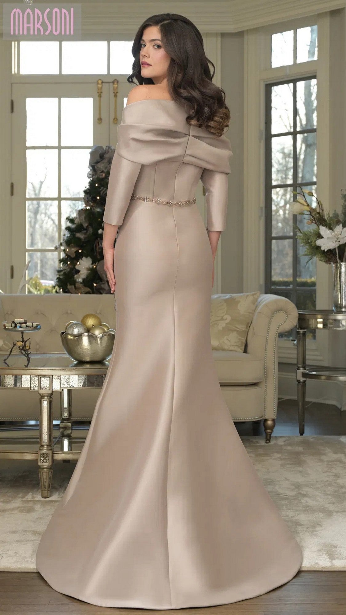 A woman in the Marsoni MV1375 long formal dress, featuring a fitted mermaid silhouette and off-shoulder neckline in taupe, stands in a stylish living room with elegant decor and large windows.