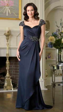 A woman in a navy Marsoni MV1377 long formal dress with beaded cap sleeves and an empire waist stands in a living room near a fireplace and floral arrangements.