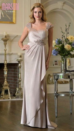 A woman poses gracefully indoors in a sand-colored Marsoni MV1377 long formal dress with beaded cap sleeves and a sparkling empire waist, standing by a fireplace and floral arrangement.
