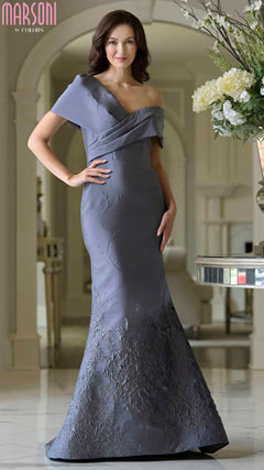 A woman poses indoors with one hand on her hip, wearing the Long Formal Dress: Marsoni MV1378 in charcoal—a fitted, asymmetric off-shoulder evening gown with textured floral details.