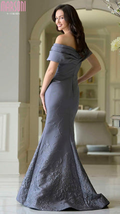 Woman stands indoors in a Marsoni MV1378 long formal dress—an asymmetric off-shoulder, floor-length grey mermaid gown with floral embroidery—smiling over her shoulder.