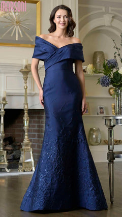 A woman in the navy Marsoni MV1378 long formal dress with an off-shoulder neckline and floral texture stands in a decorated living room.