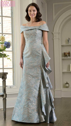 A woman in a sage Marsoni MV1381 long formal dress with an elegant off-the-shoulder neckline stands indoors near a window and floral arrangement.