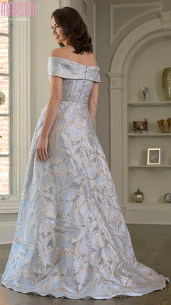 A woman wearing the Marsoni MV1382 long formal dress in light blue with a floral pattern stands in a well-lit, elegant room, facing away from the camera.