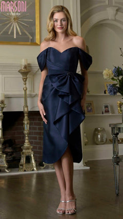 A woman stands indoors in front of a fireplace, wearing the Marsoni MV1389 knee-length semi-formal dress in navy with ruffle details and silver heels, surrounded by candlesticks and decorative items.